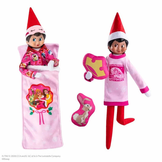 A Disney Elf on the Shelf doll shown in two outfit configurations on a white background: one wrapped in a pink blanket with a lollipop graphic, and one standing in a pink pajama set with a puzzle-piece hat and bunny-shaped accessory.