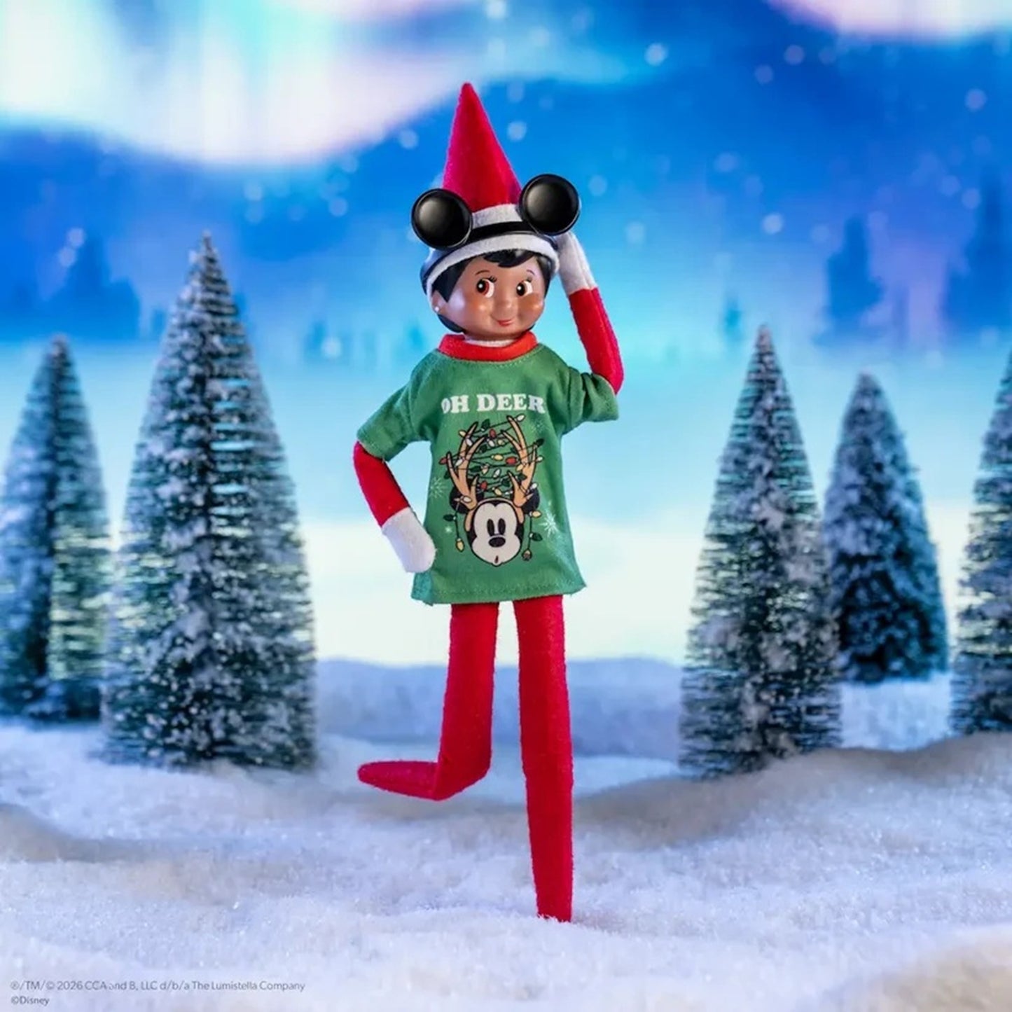Elf on the Shelf character wearing a green Mickey Mouse Christmas shirt with 'OH DEER' text, red pants, white gloves, and a red hat with black ears, posed in a snowy outdoor scene with pine trees and a blue winter background.