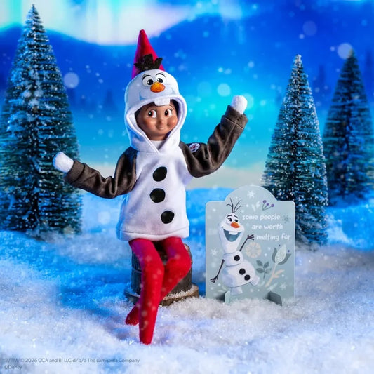 A person dressed as Olaf the snowman stands in a snowy miniature scene with small evergreen trees and a tombstone sign. The costume includes a white hooded snowman face, brown sleeves, red pants, and a red hat, set against a blue winter backdrop.