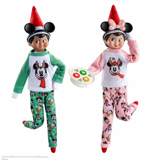 Two children's holiday pajama sets shown on doll-like mannequins against a white background. One set is green with a white chest panel featuring a cartoon face and red scarf motif, paired with green patterned pants.