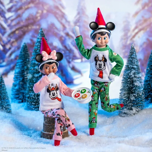 Two Disney-themed children's holiday pajama dolls posed in a snowy miniature scene with pine trees. One doll wears a white and green Santa-themed long-sleeve pajama set with green patterned pants, a red hat, and red shoes.