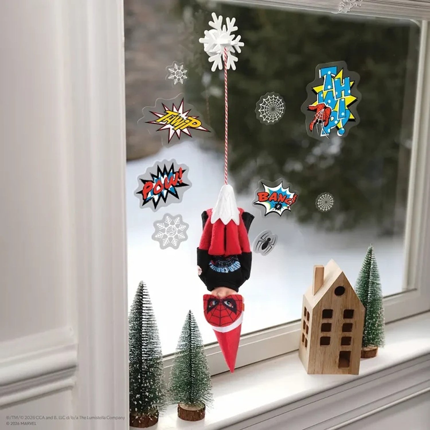 A festive Spider-Man themed window hanging decoration featuring a hanging plush figure of Spider-Man upside down, surrounded by comic-style speech bubble stickers and snowflake decals. The scene is set on a windowsill with miniature Christmas trees and a small wooden house, looking out onto a snowy outdoor background.