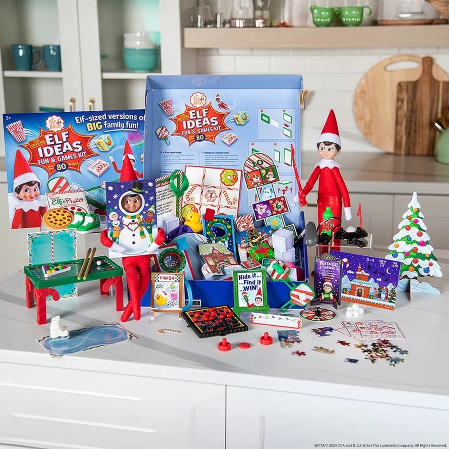 A Christmas-themed elf activity kit displayed on a white kitchen counter, featuring an open blue box, multiple colorful game components, cards, small furniture pieces, candy-themed items, and two Elf on the Shelf figures in red and white outfits. The background shows a blurred kitchen interior.