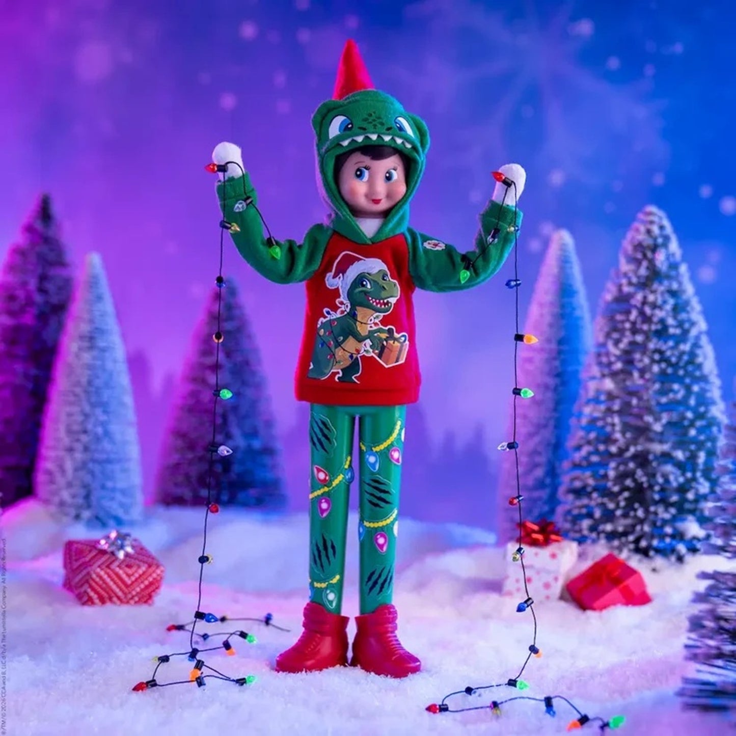 A doll or toy figure dressed in a Christmas dinosaur-themed holiday outfit, standing in a snowy miniature scene with artificial Christmas trees, wrapped gifts, and hanging string lights.