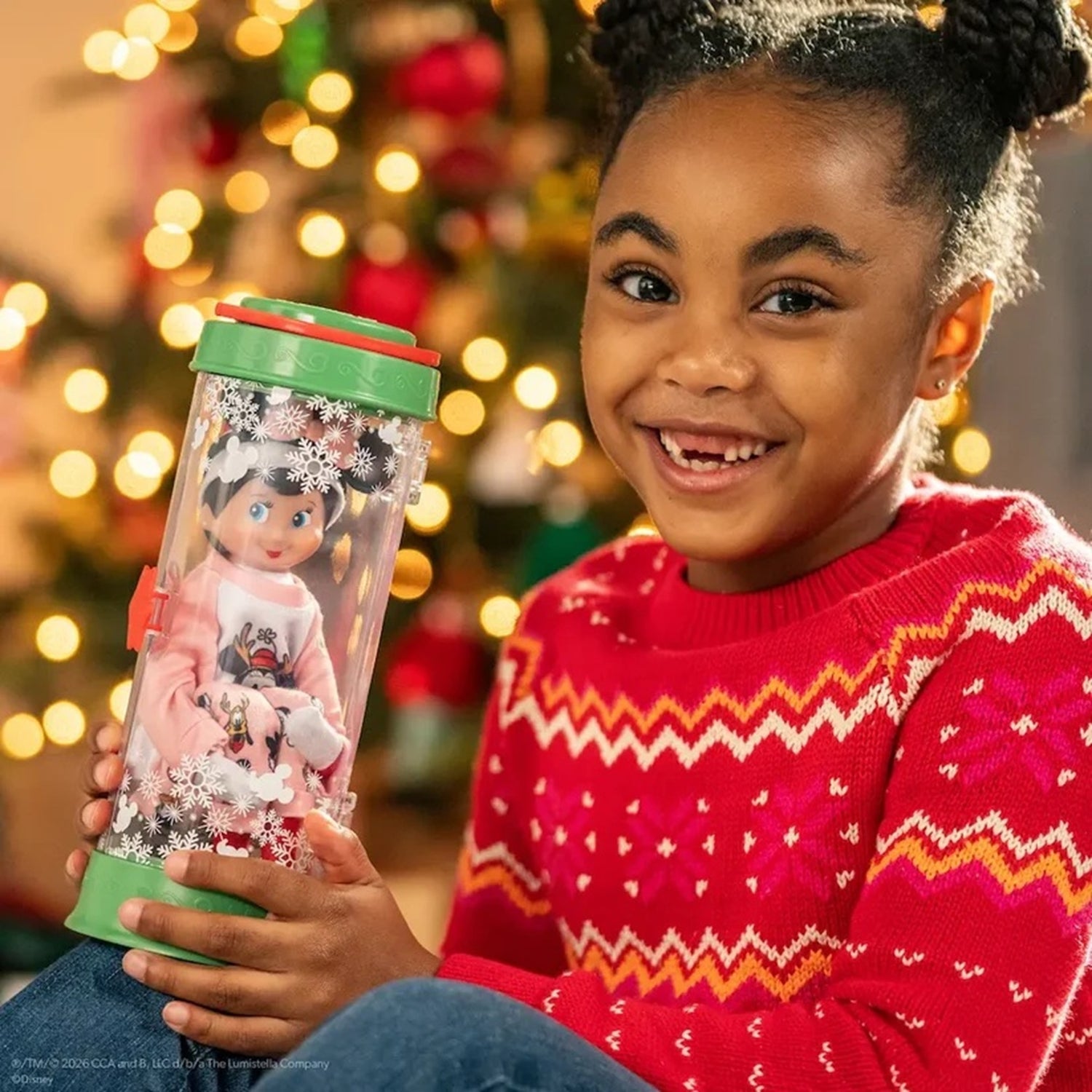 A young girl with her hair in two buns smiles while holding a clear cylindrical ornament container with a green lid. The container holds a doll dressed in pink with snowflake decorations.