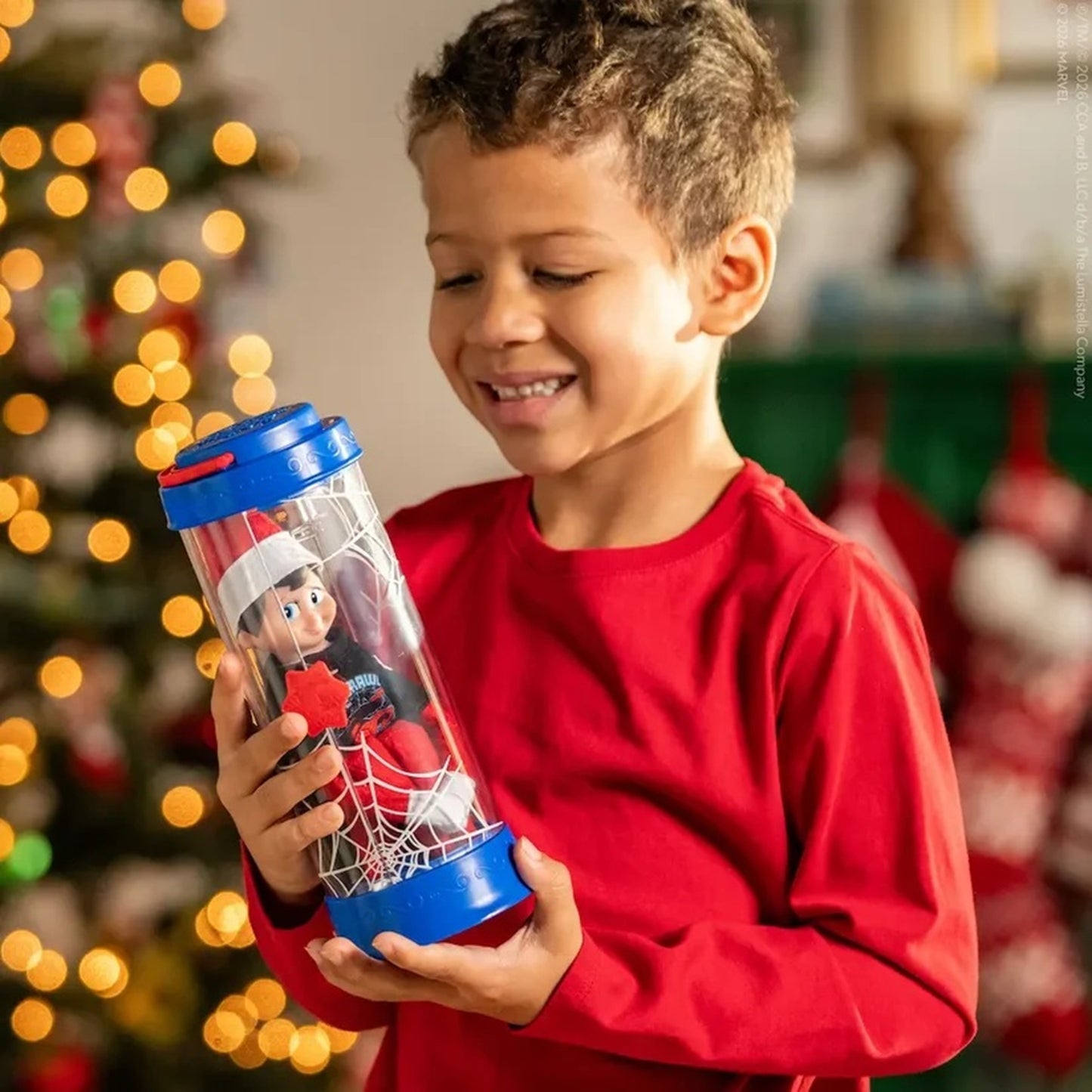 A young boy in a red long-sleeve shirt holds a clear plastic Spider-Man themed tumbler with blue lids and a Spider-Man figure inside, set against a festive Christmas background with warm bokeh lights.