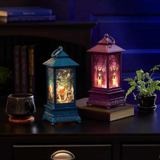Two decorative lantern-style table lamps with warm glowing lights sit on a dark wooden surface in a dimly lit room. One lantern is teal-blue and the other is purple, both with ornate carved sides and fairy-like illuminated figures inside.