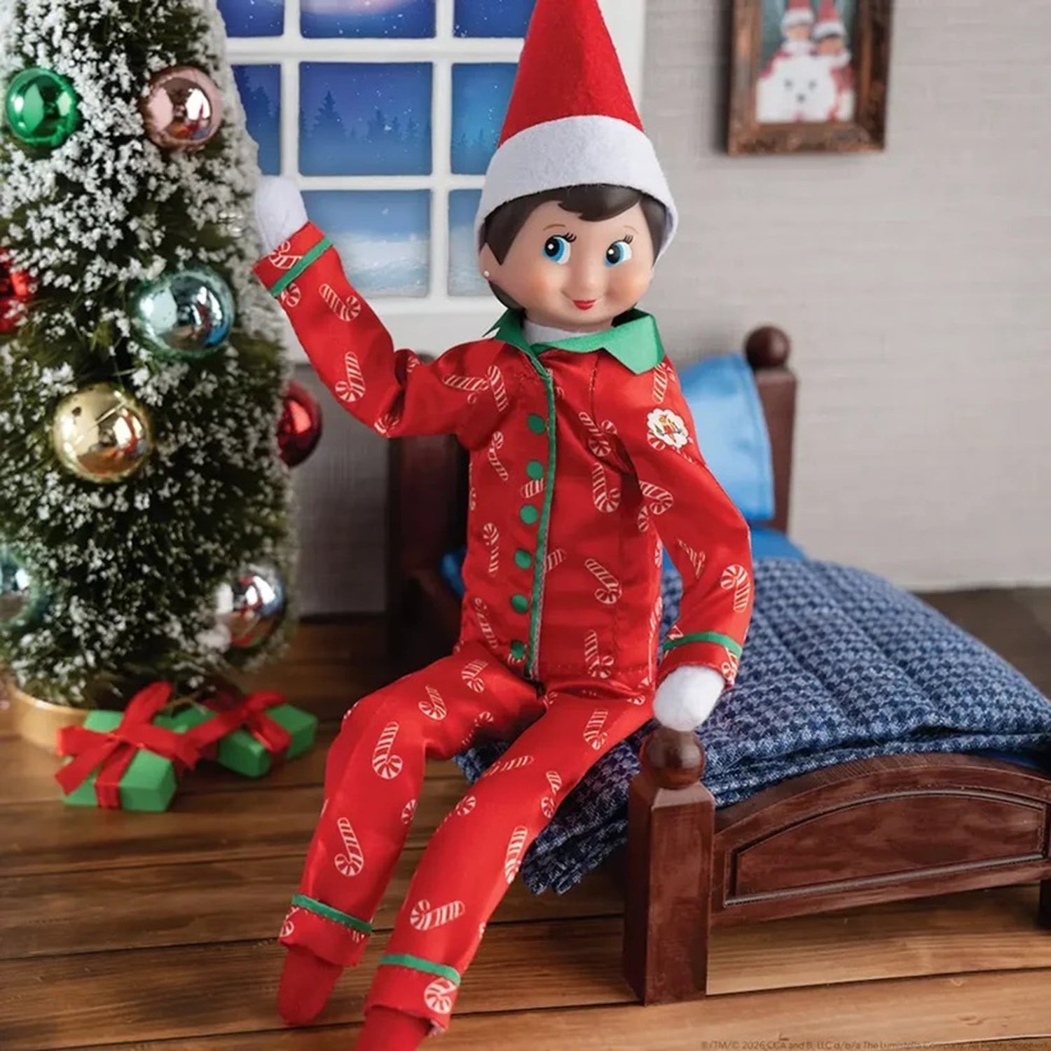 Elf on the Shelf in a red outfit with candy cane pattern sitting on a bed next to a Christmas tree.