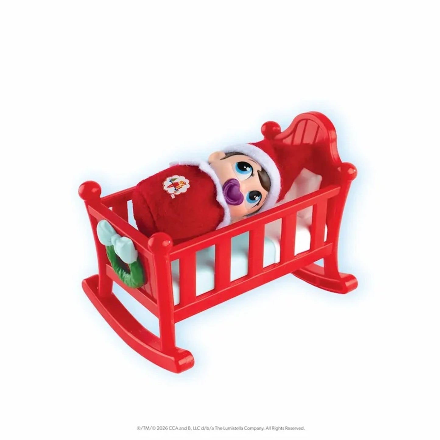 A small plush doll dressed in a red Santa outfit and hat, lying inside a red rocking cradle on a plain white background.
