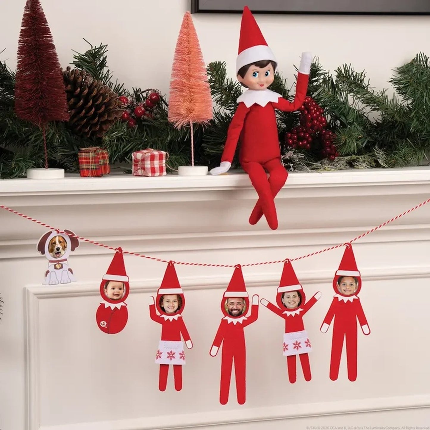 A Christmas-themed nursery or playroom scene with a red elf costume garland hanging below a mantel, alongside a red elf doll sitting on the mantel. The garland includes multiple red-and-white elf cutouts with printed faces and a dog-shaped ornament.