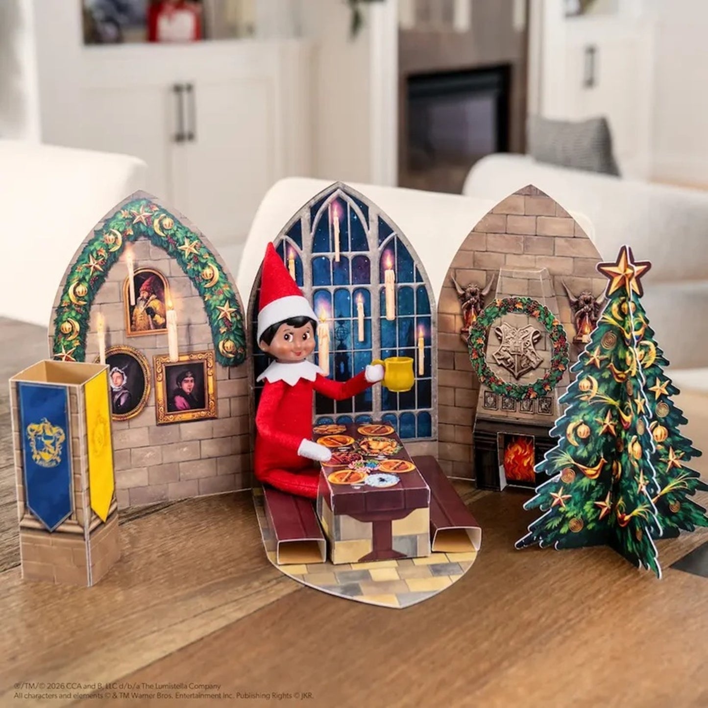 A Christmas-themed pop-up cardboard scene displayed on a wooden table in a living room. The scene features a small Elf on the Shelf figure sitting at a table with a yellow mug, flanked by two arched windows, a fireplace with a wolf wreath, a lit Christmas tree, and a blue-and-gold banner.