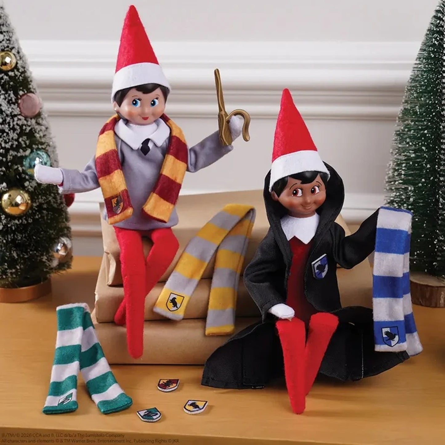 Two elf dolls dressed as Slytherin and Harry Potter, posed on a wooden table with their scarves and accessories. The background features a white paneled wall and miniature Christmas trees.