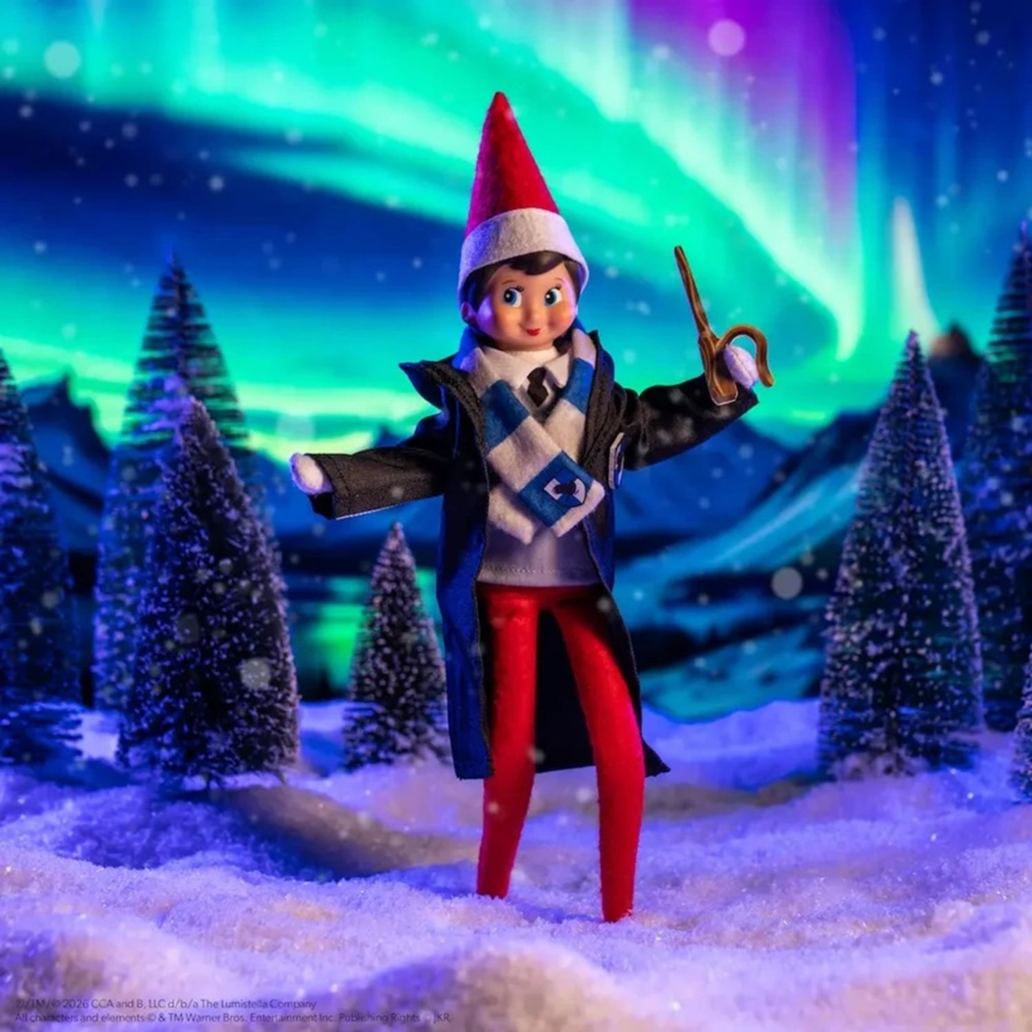 A toy elf doll dressed in a wizard outfit with a red Santa hat, dark coat, blue scarf, and red pants, standing in a snowy miniature scene with glowing aurora lights in the background.