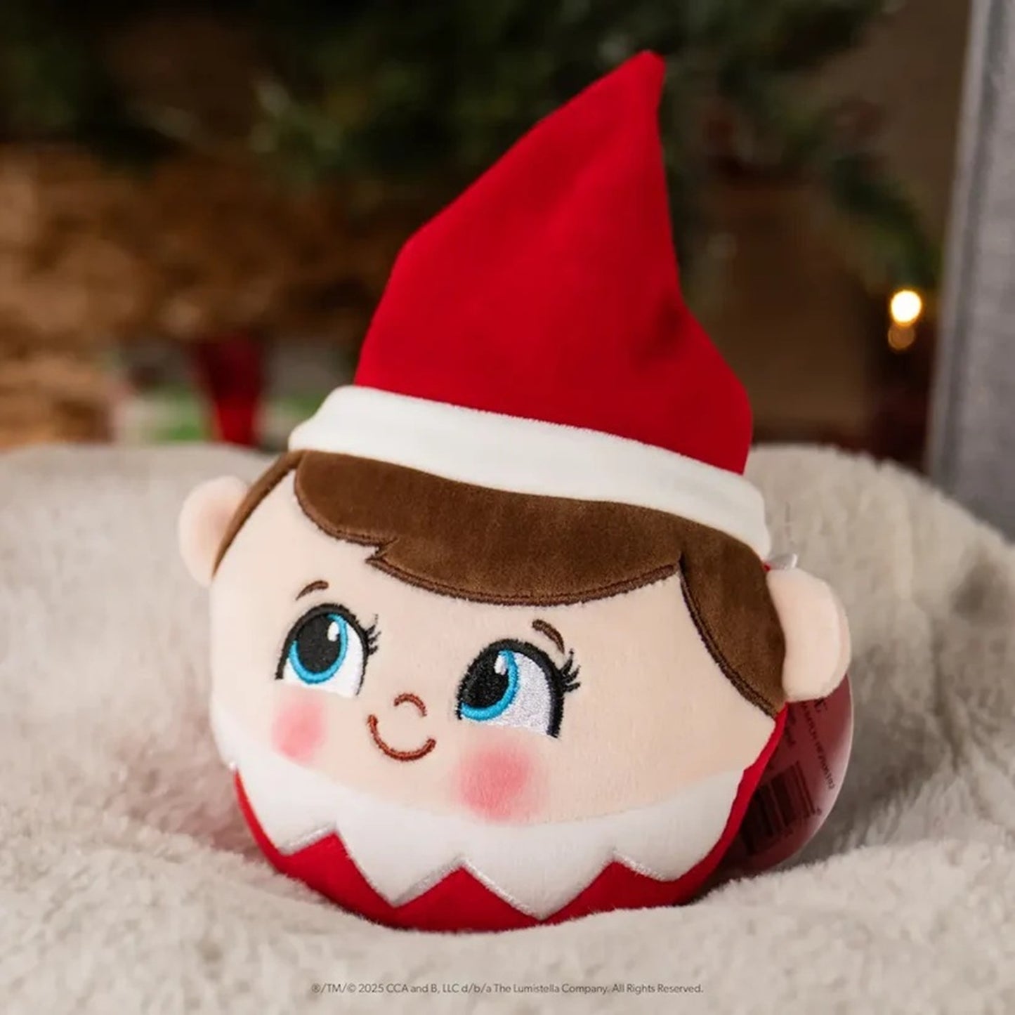 A plush Elf on the Shelf toy with embroidered facial features, blue eyes, pink cheeks, brown hair, and a red hat, resting on a soft white blanket with a blurred Christmas tree in the background.