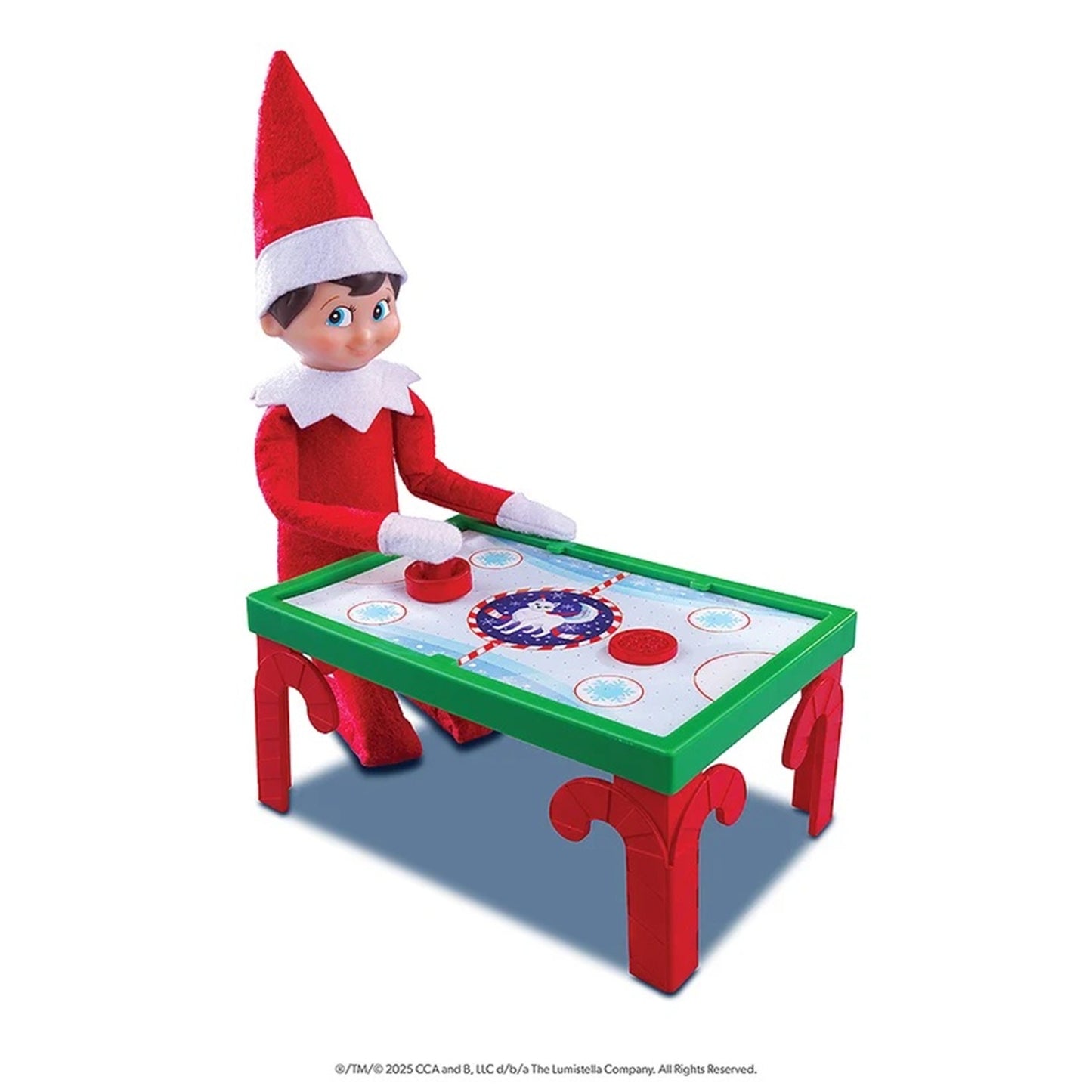 A plush Elf on the Shelf toy positioned behind a small red and green hockey table, on a plain white background.