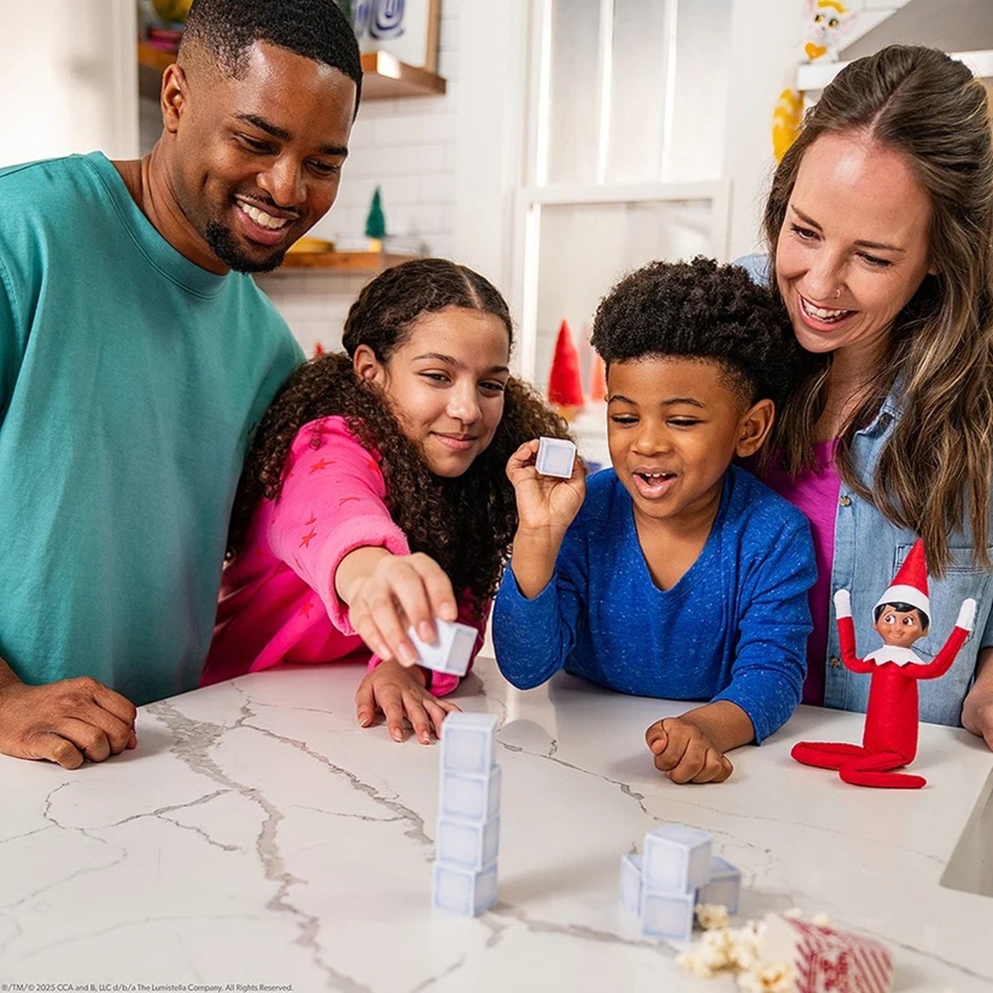 A family of four smiles and plays with white building blocks on a white marble kitchen countertop, with a small red Elf on the Shelf toy sitting on the counter.