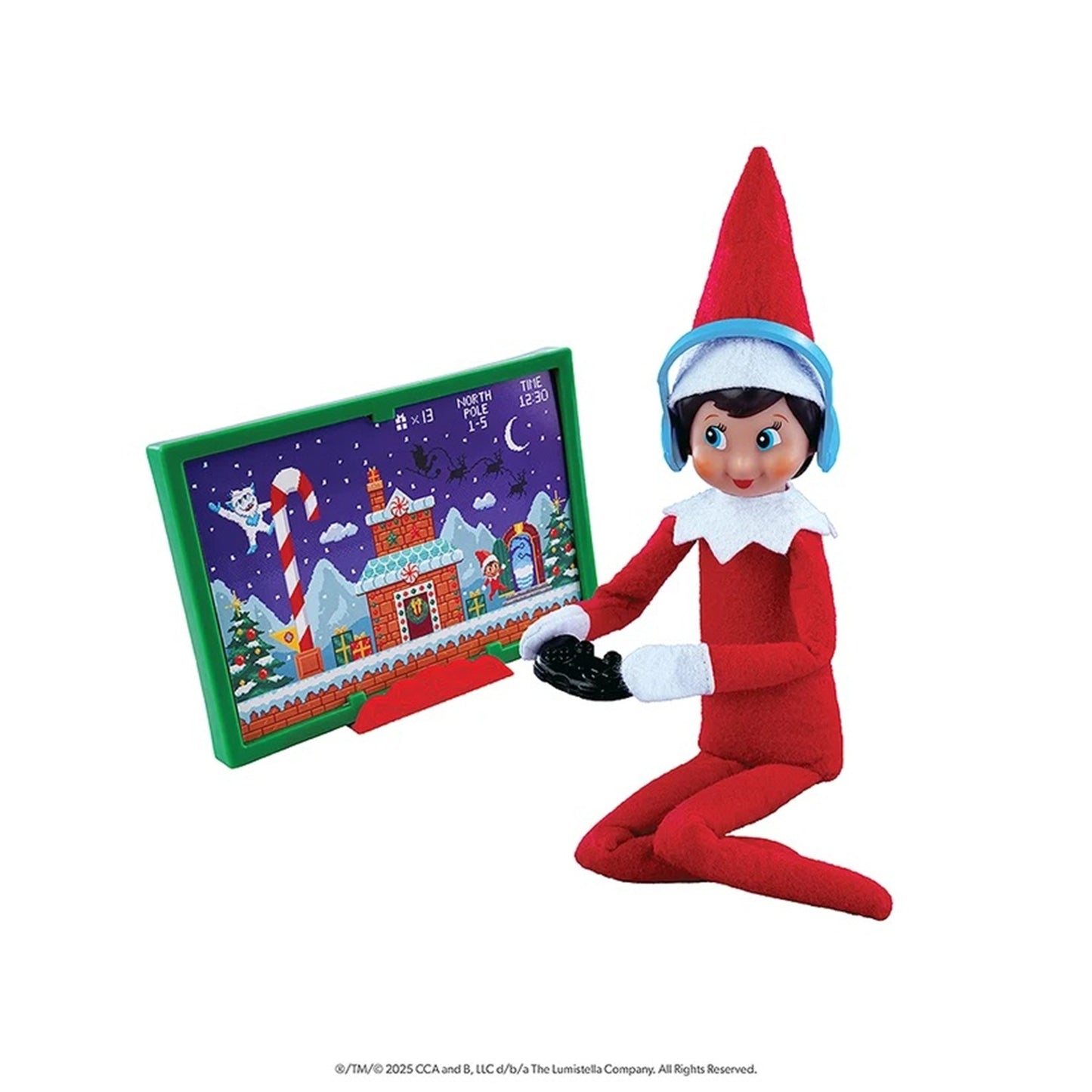 Elf on the Shelf toy figure kneeling beside a green-framed picture frame showing a Christmas village scene, on a plain white background.