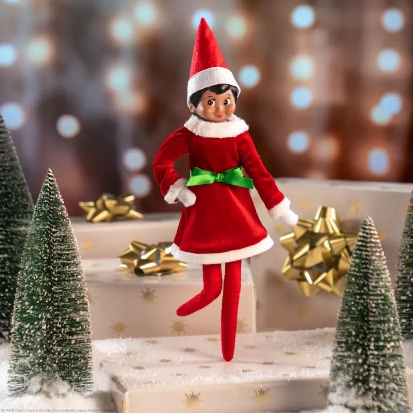 Elf on the Shelf figurine in a red outfit with a green bow, standing on a decorative Christmas scene with trees and presents.